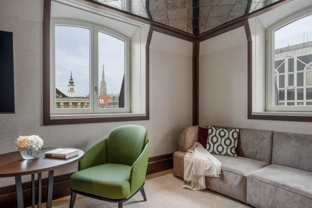 Ein modernes Hotelzimmer im Almanac Palais Vienna verfügt über einen grünen Stuhl, ein beiges Sofa, einen Beistelltisch mit einem Buch und Blumen sowie große Fenster mit Blick auf die Gebäude der Stadt und die Kirchtürme.